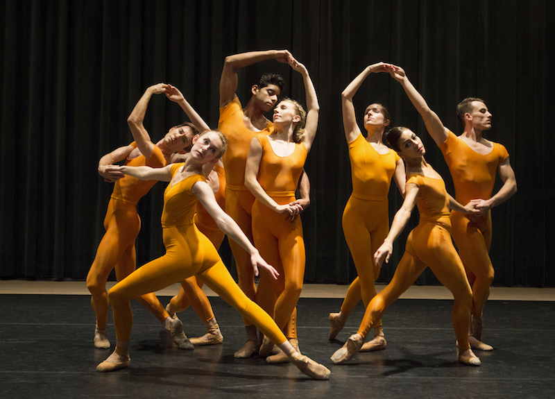 A group of dancers link hands to form a human chain in marigold unitards